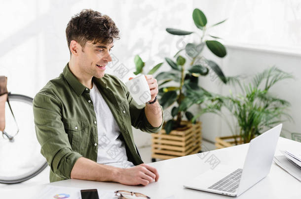 穿着绿色衬衫的英俊商人坐在桌旁, 带着笔记本电脑, 在办公室喝咖啡