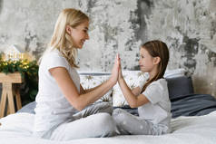 照片上的单身母亲和女儿一起玩耍，穿着睡衣坐在床上，做五步姿势，在家里休息。妈妈和小女孩拍手叫好，面带微笑