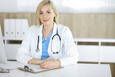 在诊所的办公桌前工作的女医生的肖像。勃隆德快乐的医生填写医疗表格或处方
