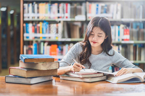 亚洲青年学生穿着休闲服在大学图书馆阅读和做作业或与各种书籍和固定在书架背景的木桌上回到学校