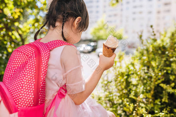 可爱的小女孩坐在城市街道上, 在户外吃冰淇淋的后视图。快乐的女孩在外面上课后放松。快乐的学龄前儿童与背包。人、教育