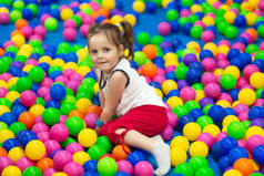漂亮的小女孩在娱乐商城的儿童区玩耍, 躺在五颜六色的球上, 正面微笑, 看着相机。小孩子用两匹小马尾巴玩彩色球。童年概念