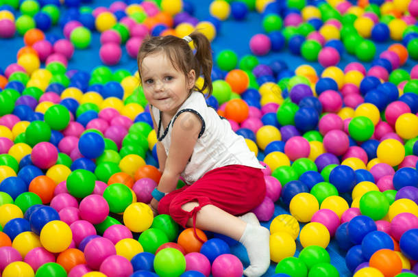 漂亮的小女孩在娱乐<strong>商城</strong>的儿童区玩耍, 躺在五颜六色的球上, 正面<strong>微</strong>笑, 看着相机。小孩子用两匹小马尾巴玩彩色球。童年概念