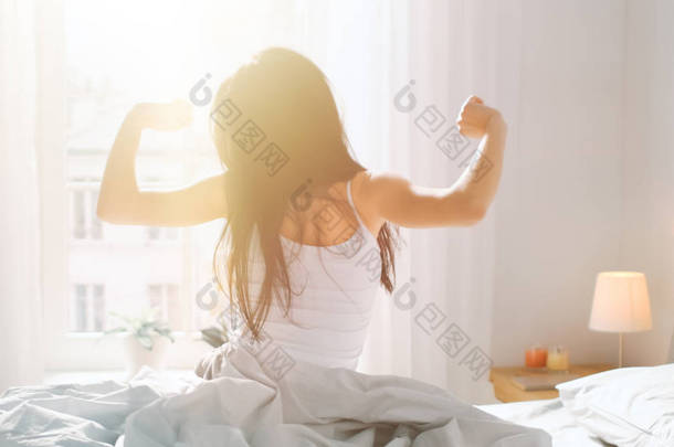 美丽的布鲁内特在早晨醒来，在床上伸展，阳光从大窗户照在她身上。<strong>快乐</strong>的小女孩，带着温暖的阳光，迎接新的一天.