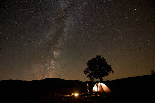男子坐在旅游帐篷和三脚架相机前燃烧营火, <strong>观看</strong>美丽的黑暗星空与银河星座