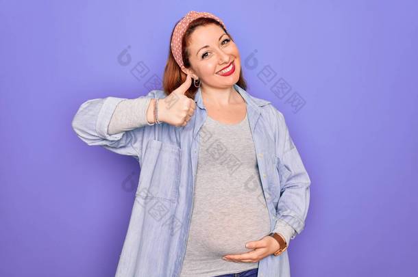年轻美丽的红头发孕妇期待着宝宝在孤立的紫色背景上做着快乐的大拇指举手表决。在镜头前看着成功的表情.