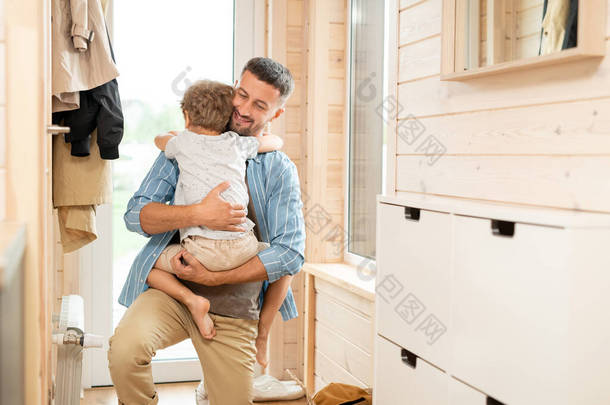 <strong>快乐</strong>的、充满爱心的年轻父亲下班回家后，单膝跪在走廊里拥抱他可爱的小儿子