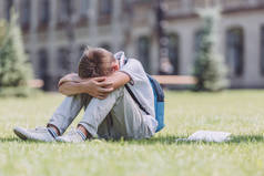 疲惫的小学生背着背包坐在学校附近的绿色草坪上
