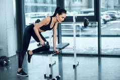 年轻运动的妇女举哑铃, 当倾斜在健身房锻炼长凳