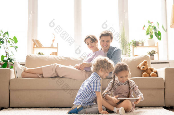 年轻快乐的父母坐在沙发上看着他们的孩子们坐在地板上在触摸板上打滚