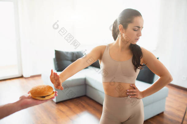 在检疫期间，年轻女子在房间里做瑜伽锻炼。女孩拒绝吃汉堡包。对不健康食物说不.
