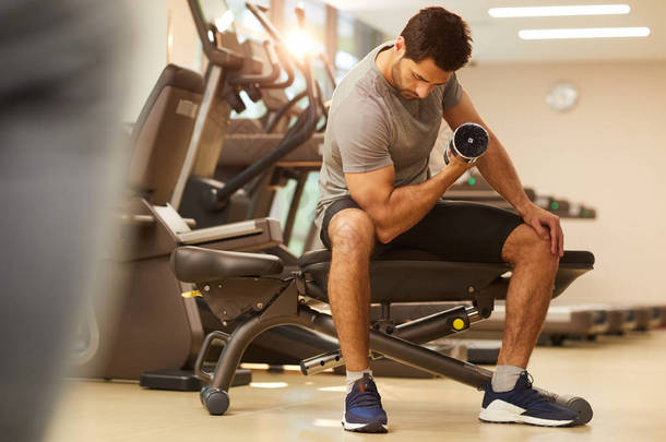 在健身房进行力量锻炼时，肌肉男子举起哑铃坐在长凳上的全长肖像，复制空间