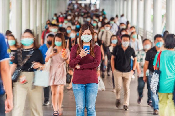 在曼谷的交通高峰时段，亚洲妇女戴着外科口罩，在一群模糊不清、难以辨认的商人中间使用手机，以防止考拉病毒在繁忙的工作日爆发