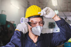 一名戴手套的男工和一名戴着口罩和硬礼帽的扫描温度工人在进入工厂前，为工厂工人保护COVID 19病毒.