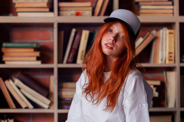 一个红头发女人的画像，她穿着<strong>模版</strong>的空白T恤和有健康雀斑皮肤的帽子。年轻的高加索模特在书架上摆姿势.复制空间和模拟。刊登广告的地方