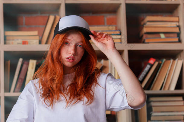 一个红头发女人的画像，她穿着模版的空白T恤和有健康雀斑皮肤的帽子。年轻的高加索模特在书架上摆姿势.复制空间和模拟。刊登广告的地方