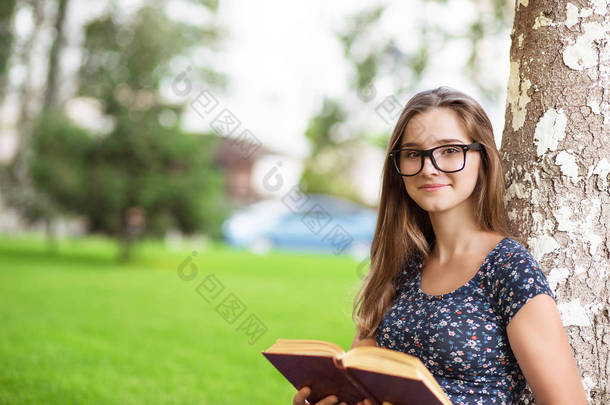 女学生穿着便服和眼镜捧着一本书, 看着你微笑着坐在校园外的绿色草坪树上的背景。多元文化模式, 混合种族, 亚洲俄罗斯女孩