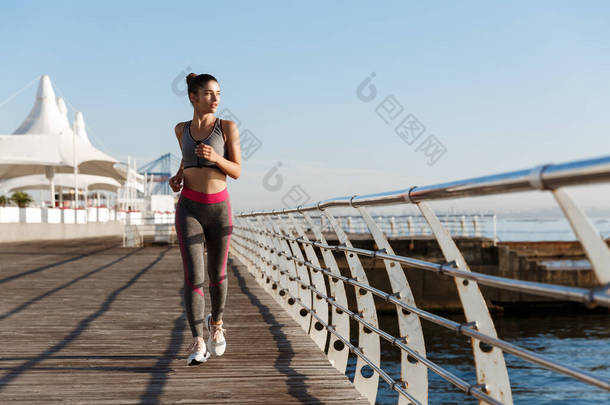全长的迷<strong>人</strong>的年轻女子沿着海滨长廊向镜头跑去，望着大海。女子赛跑选手早上在码头训练