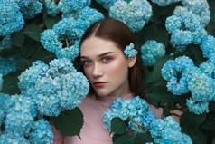 美丽的年轻女子的特写脸，红嘴唇和蓝眼睛，她的耳朵后面有一朵蓝色的花，站在花丛中