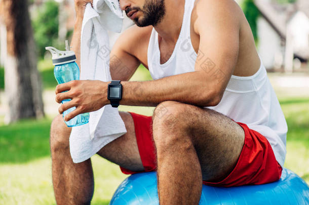 留着胡子的男子拿着运动瓶，坐在健身球和擦汗的裁剪视图