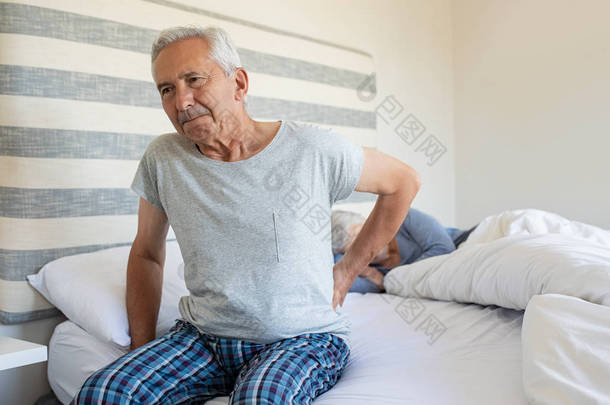 老人在家里忍受背部疼痛, 而妻子则睡在床上。有背痛的老人从床上爬起来有困难。痛苦的疼痛和坐在床上的早晨.