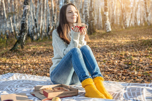 一位年轻漂亮的女士正在黄昏时分的秋天的公园里吃着披萨，喝着热茶。秋寒季节舒适生活的概念