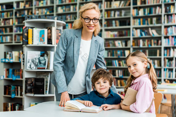 开朗的老师在眼镜站在附近的可爱的孩子在图书馆 