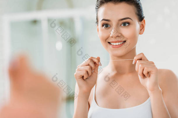 美丽的微笑的女孩拿着牙线并且看镜子在洗手间 