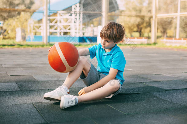 一名身穿蓝色T恤的男孩篮球运动员手拿着球坐在篮球场上，在运动后休息。体育与健康生活方式的概念