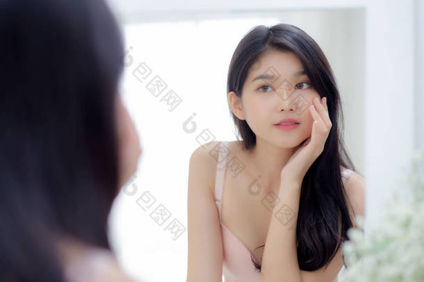 美丽的年轻亚洲女人笑着看着镜子，用皮肤护理和化妆品检查脸，以恢复青春和健康；漂亮的女孩则高兴地清洁脸，用<strong>奶油</strong>或乳液洗脸，以增进健康.