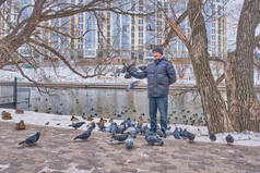 今年冬天，穿着羽绒服、牛仔裤和针织帽子的快乐的亚裔老人在河边的一个城市公园里喂鸟。他看着相机，微笑着。鸽子正坐在他的手上.