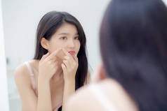 美丽的年轻亚洲女人看着镜子和触摸的脸，是一种痤疮、青春痘、美容美发、脸上皱纹、化妆品和化妆品的概念.