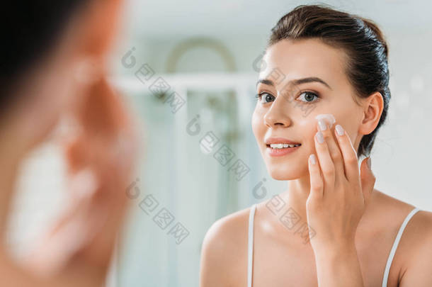微笑的年轻女子在浴室里涂抹面霜和镜子的选择焦点 