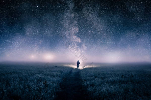 在 photoshop 中编辑的数字艺术图片, 是一个人拿着火炬在田野里的剪影, 在一个喜怒无常的雾蒙蒙的夜晚, 看着地平线上的灯光。头顶上有一颗恒星的星系. 