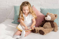 可爱的开朗的孩子玩泰迪熊和阅读书籍在床上的儿童房
