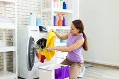 亚洲家庭主妇正在把衣服从篮子里分离出来，放进洗衣机里洗衣服。年轻女人快乐地做家务.