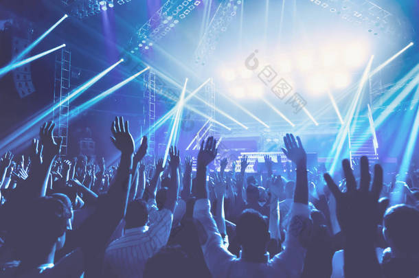 年轻人与愉快的晚上在<strong>音乐会</strong>, <strong>人群</strong>用举起的手站立在蓝色光, 唱歌和享受音乐概念