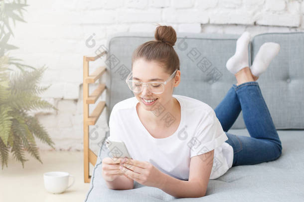 漂亮的欧洲白种女孩的水平图片躺在灰色沙发上有塑料透明眼镜手持手机, 与朋友聊天或浏览<strong>移动互联网</strong>