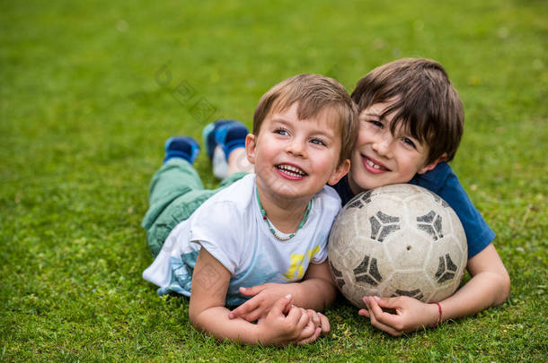 两个孩子抱着足球躺在草地上，踢足球后笑着放松
