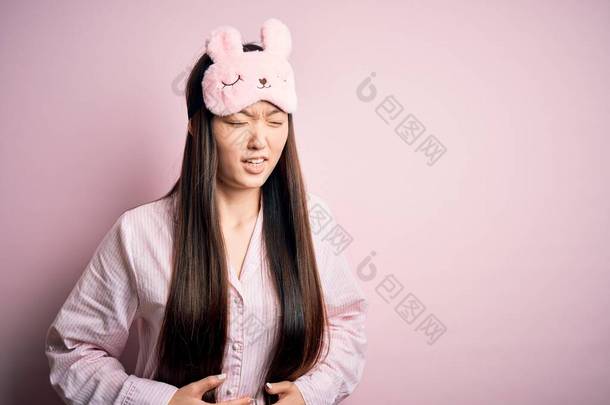 年轻的亚洲女人，头戴睡衣，戴着睡眠面罩，带着粉色的隔离背景，双手放在肚子上，因为恶心、痛苦的疾病感觉不舒服。阿切概念.
