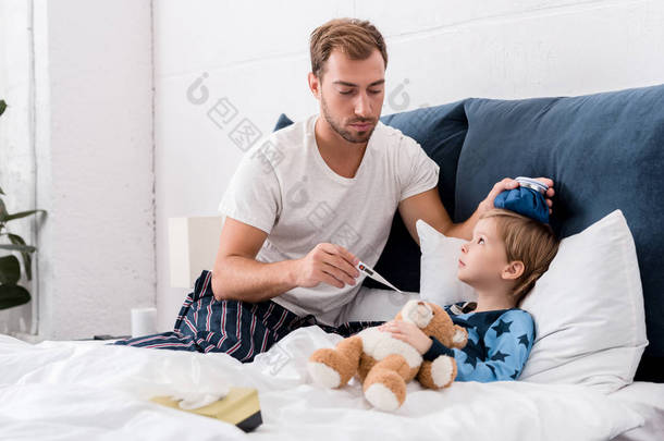 父亲检查儿子体温与电温度计和在他的头在床上举行冰包