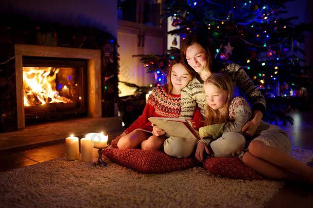 圣诞节前夕，妈妈和她的两个可爱的小女儿在温暖舒适的客厅里用平板电脑在家里壁炉边.