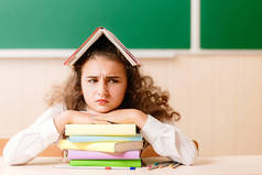 女生在学校制服坐在她的办公桌上的书籍和铅笔的背景下的绿色板。那女孩用一本书遮住了她的头。