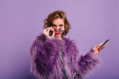 感兴趣的女孩在时尚的紫色毛皮大衣期待相机拿着闪闪发光的太阳镜。自信的女模特的Indor肖像与智能手机在明亮的背景上摆姿势..
