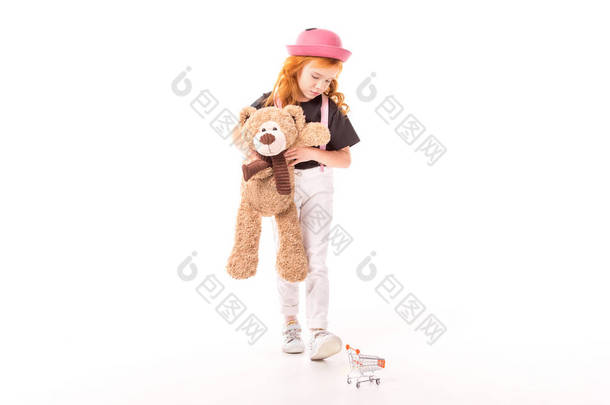 心烦意乱的孩子抱着泰迪熊, 看着在白色的购物车玩具