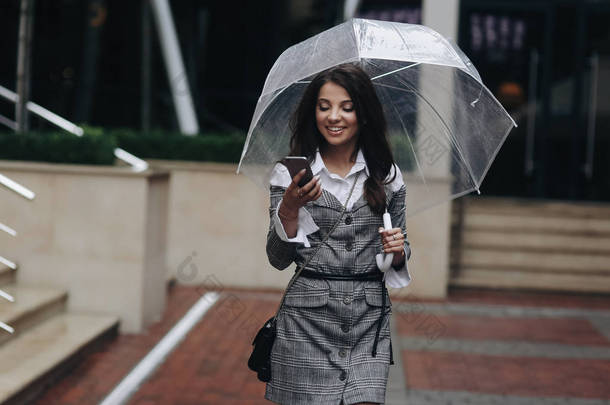 美丽的女人的肖像与智能手机在手和打字短信站在办公室附近。在伞下穿灰色外套的女人收到一个有趣的短信