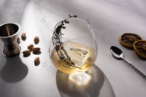 透明玻璃杯，香草，冰块和威士忌，白色桌子上的阴影靠近开心果，勺子，干柑橘片，吉格