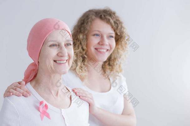 积极老年母亲患有红斑狼疮戴着粉红色的头巾和丝带,女儿站在她身后支持她