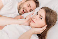 不满的女人靠在打鼾的丈夫身边躺在床上，用手塞住耳朵，看着摄像机