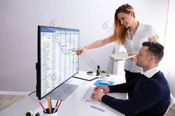 微笑的女商人指着电脑屏幕，而商人分析甘特图表在工作场所